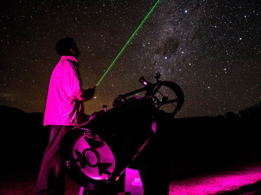 Guía señalando el cielo con un láser junto a un telescopio bajo la Vía Láctea